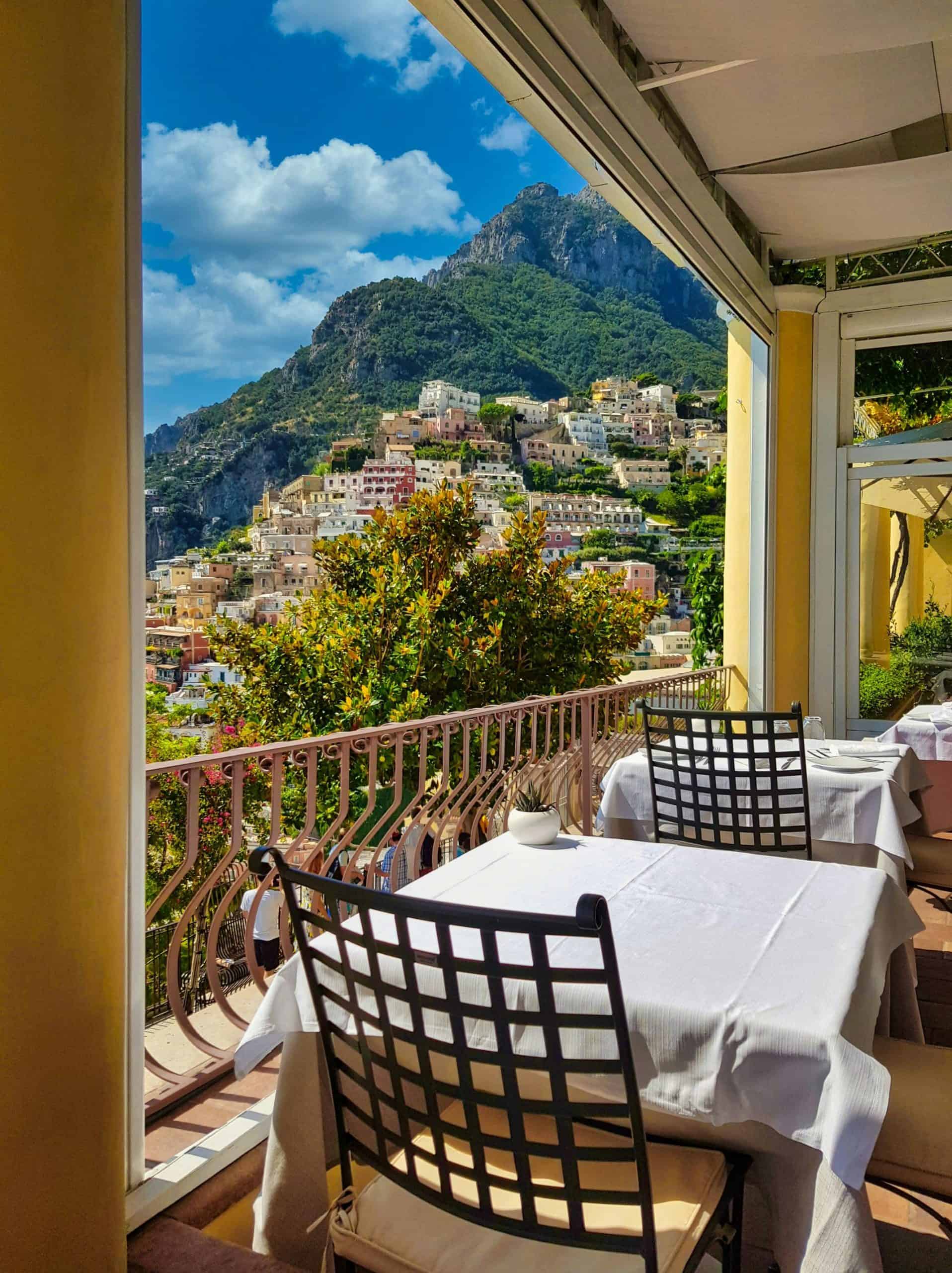 A table and chairs overlooking the view of buildings cliffside with the mountains in  the background