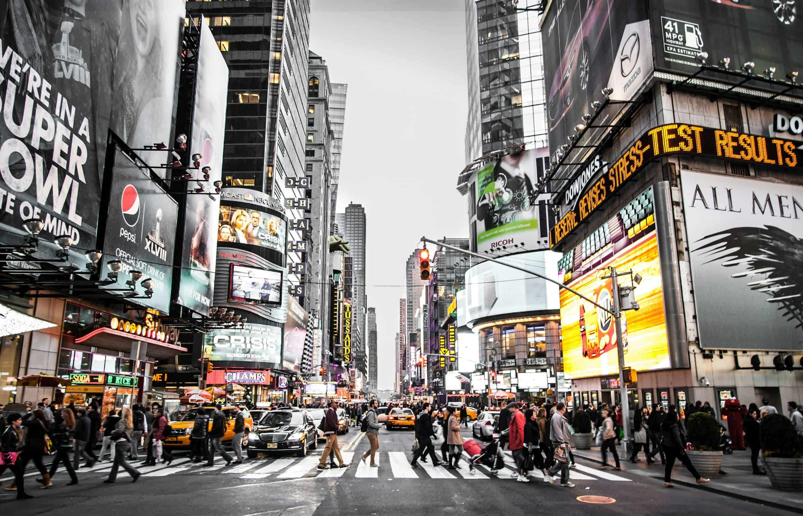 A crowd of people crossing the street in New York near Times Square with large digital billboards and buildings in the background