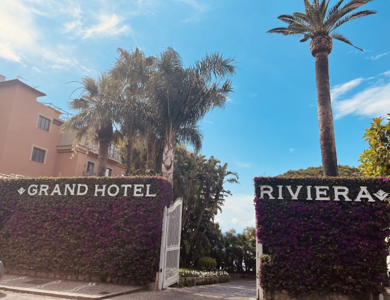 Gated entrance covered in flowers with the words Grand Hotel Riviera and palm trees in the background