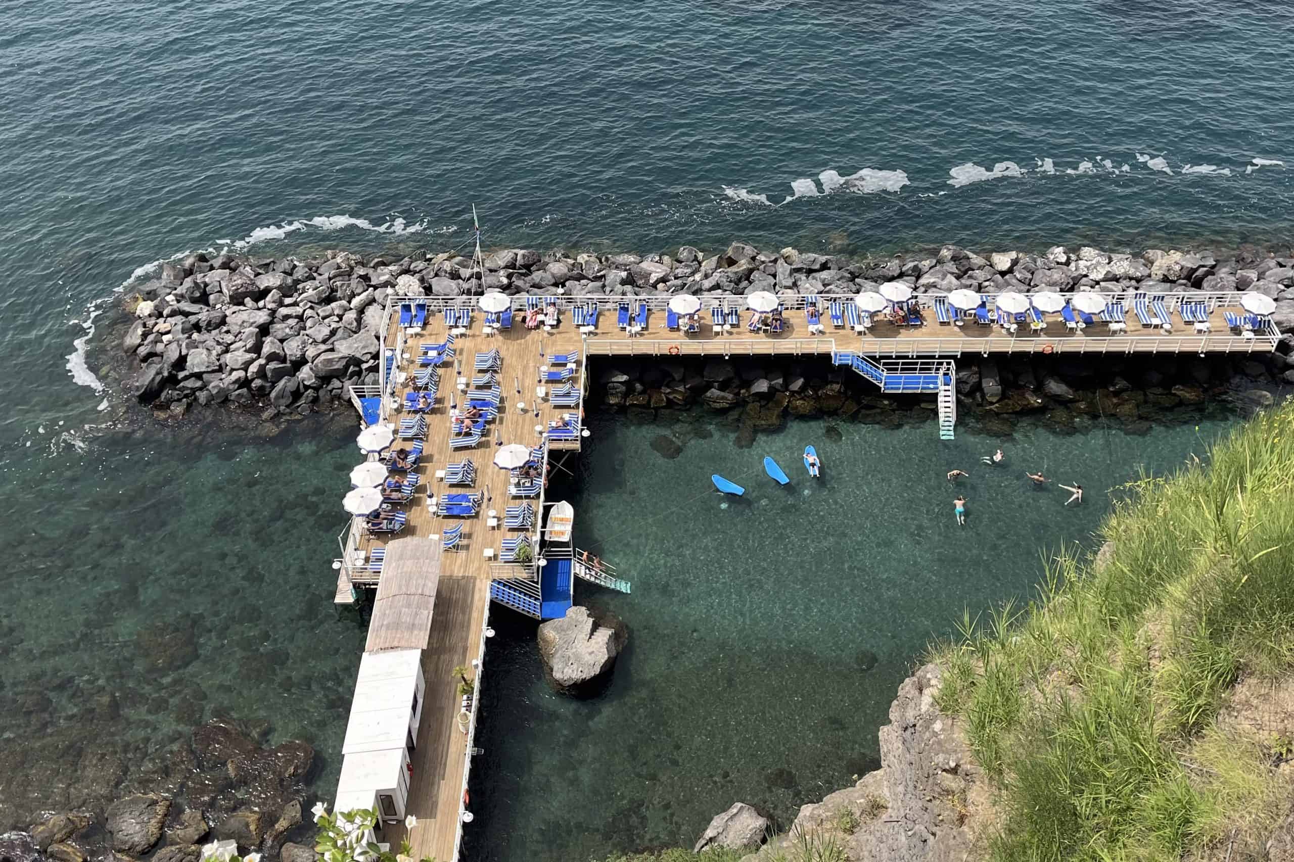 Aerial view of a sun deck platform with blue and white striped chairs and white umbrellas above the sea and people swimming in the water