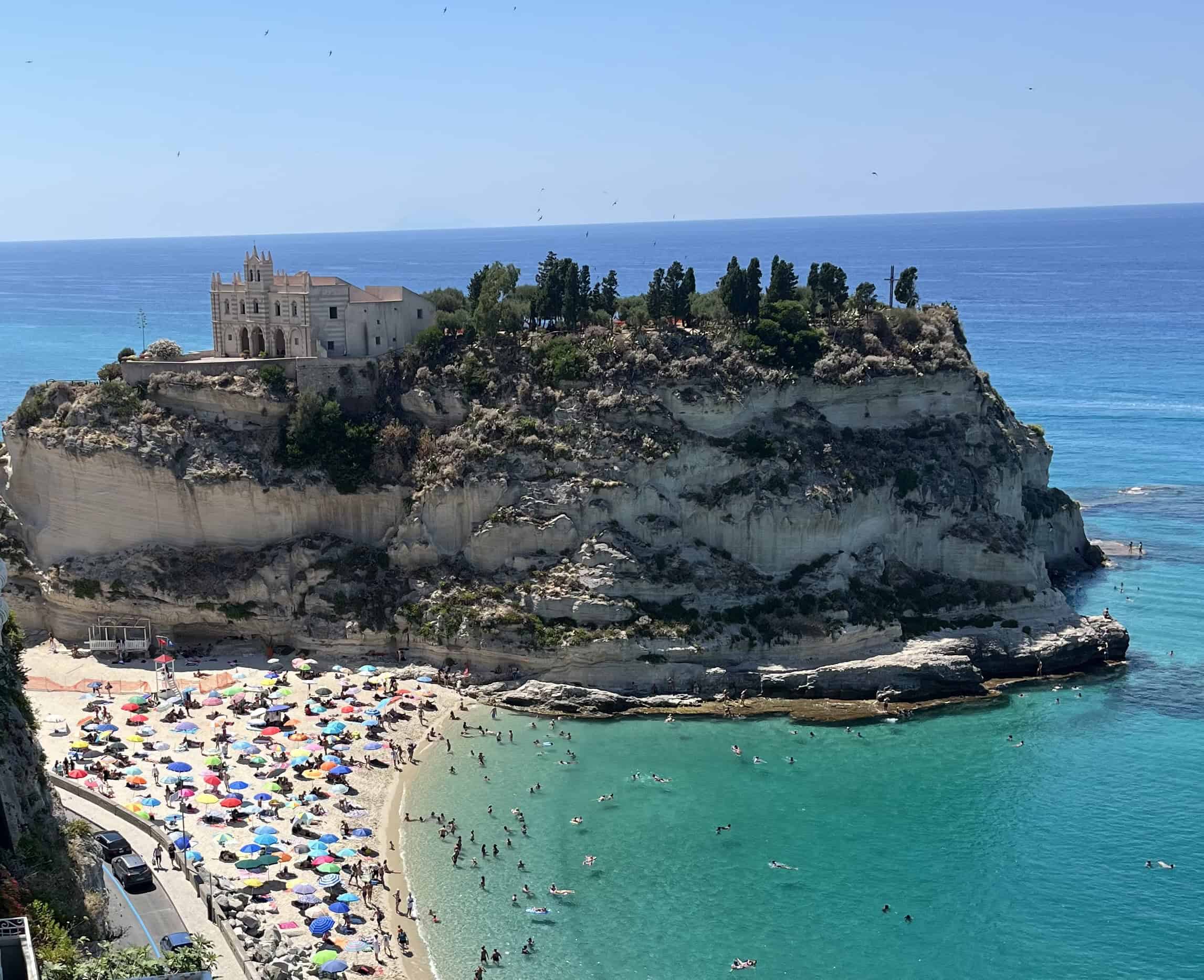 Beach with umbellas and people swimming in the water next to an island with a building on top of it