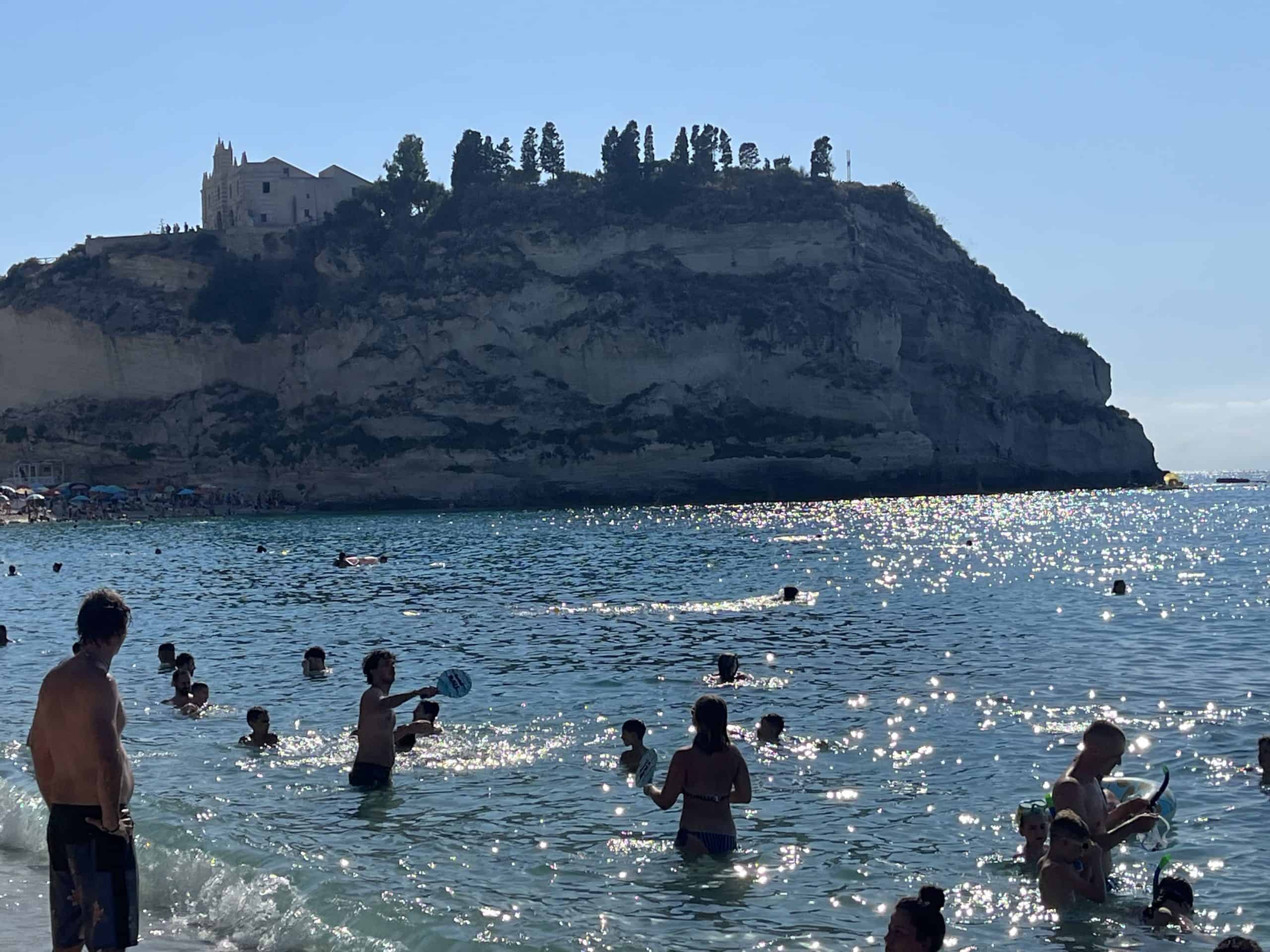 People swimming in the water with the Santa Maria Sanctuary on a cliff in Tropea in the background