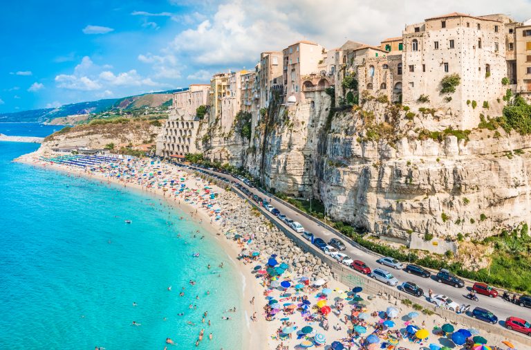 Buildings on a cliff next to a winding road with cars next to a beach and ocean blue sea