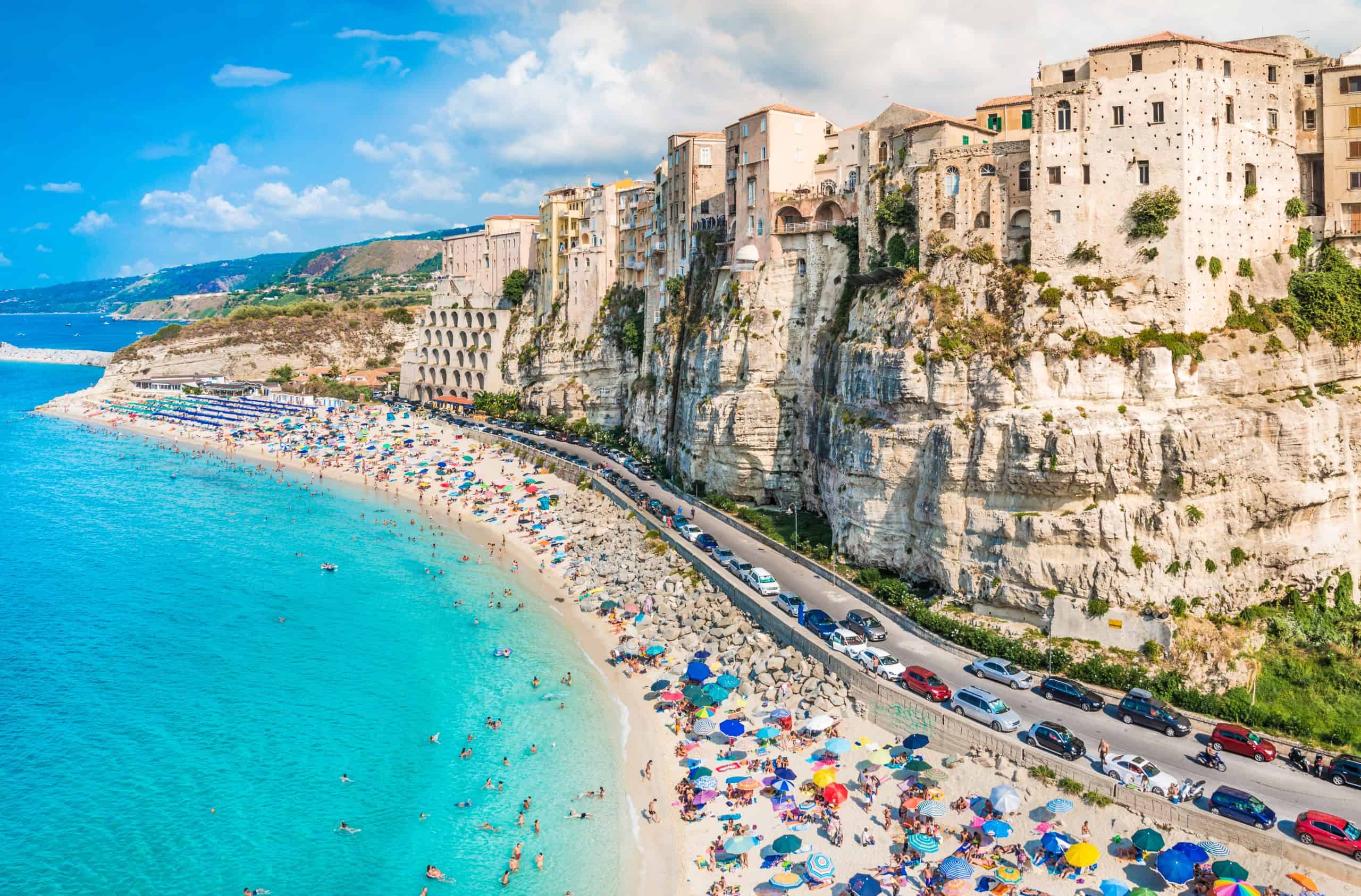 Buildings on a cliff next to a winding road with cars next to a beach and ocean blue sea