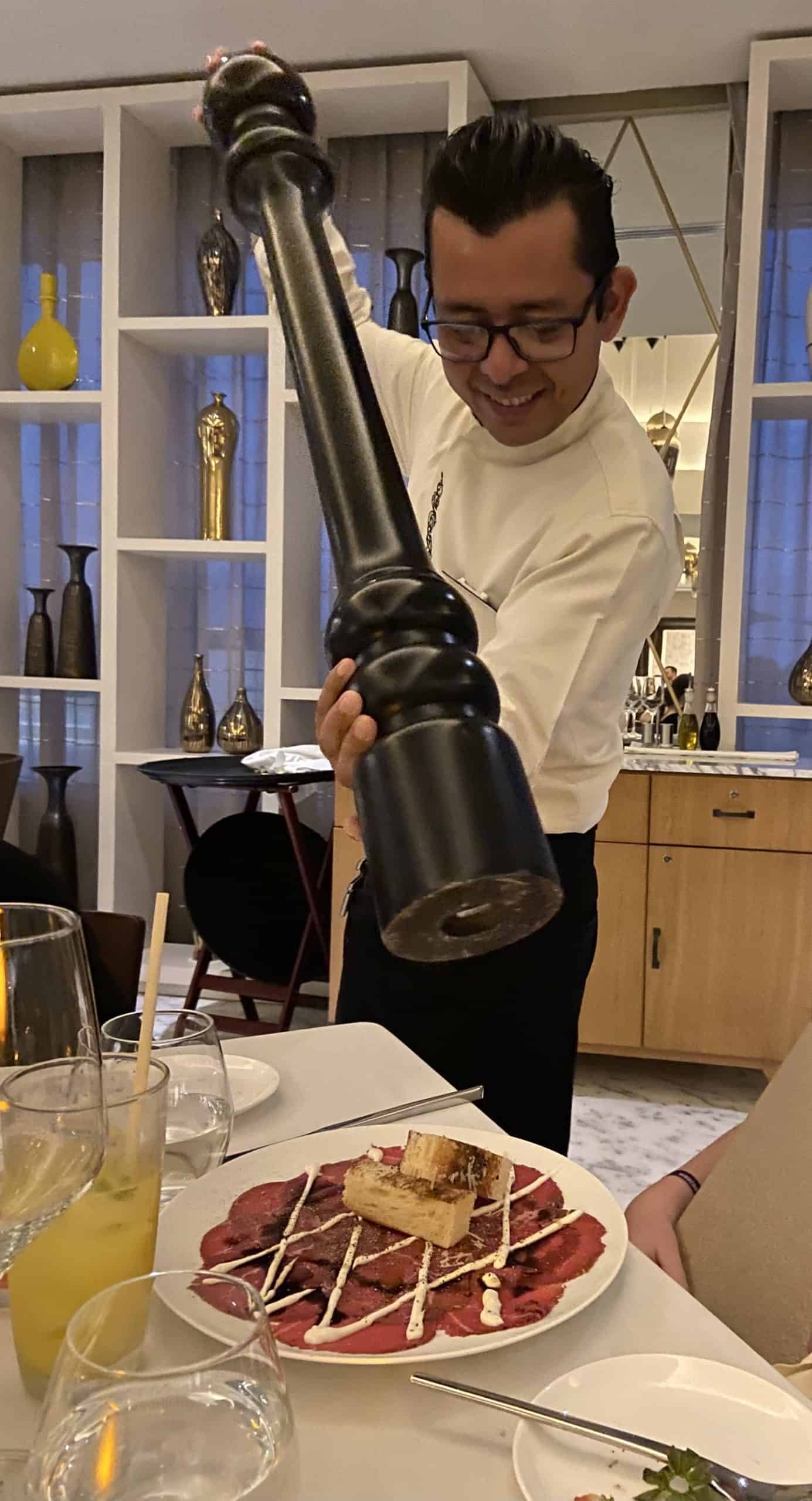 a man holding a giant pepper shaker over a plate of beef slices