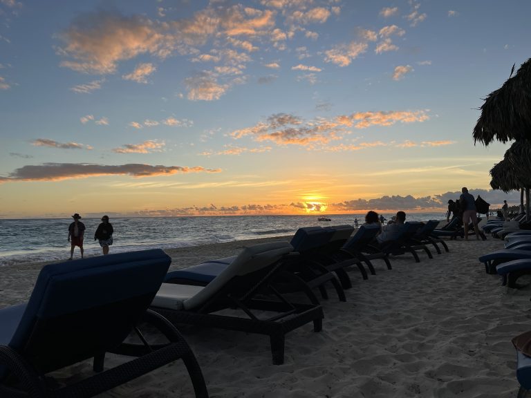 Orange sun rising white clouds in light blue sky people walking on beach and beach chairs