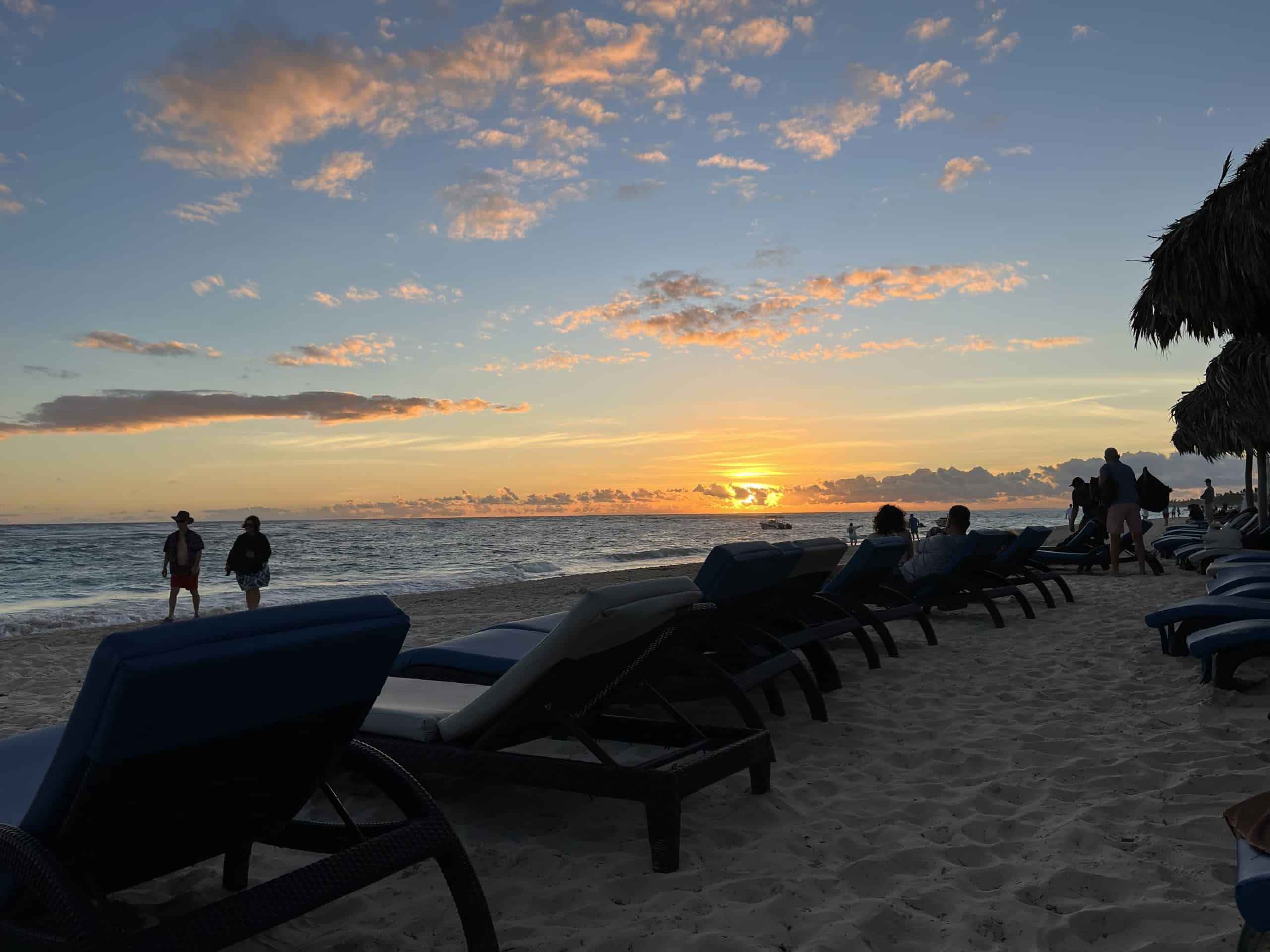Orange sun rising white clouds in light blue sky people walking on beach and beach chairs