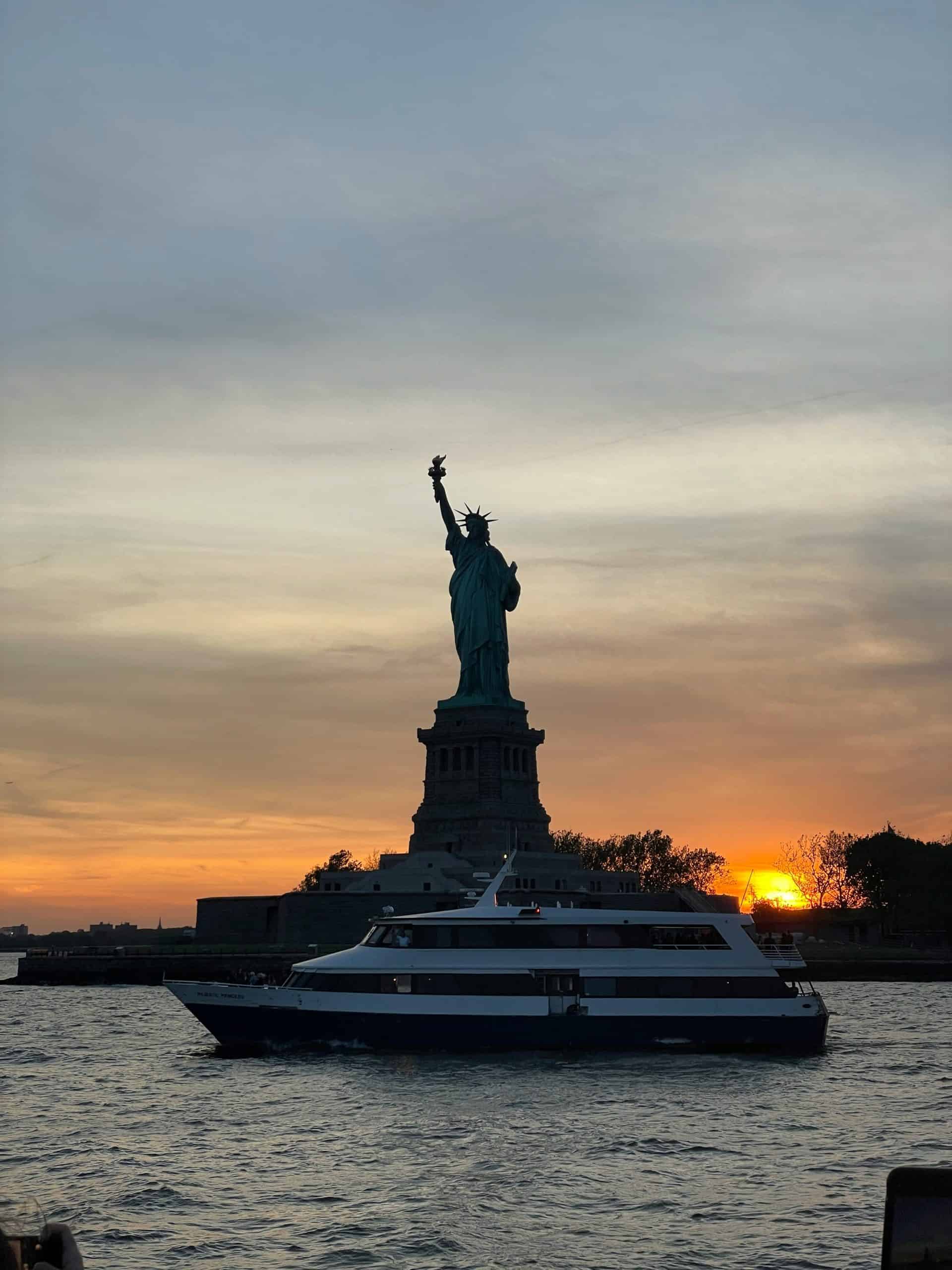 sun setting behind the Statue of Liberty with a boat in front on the water