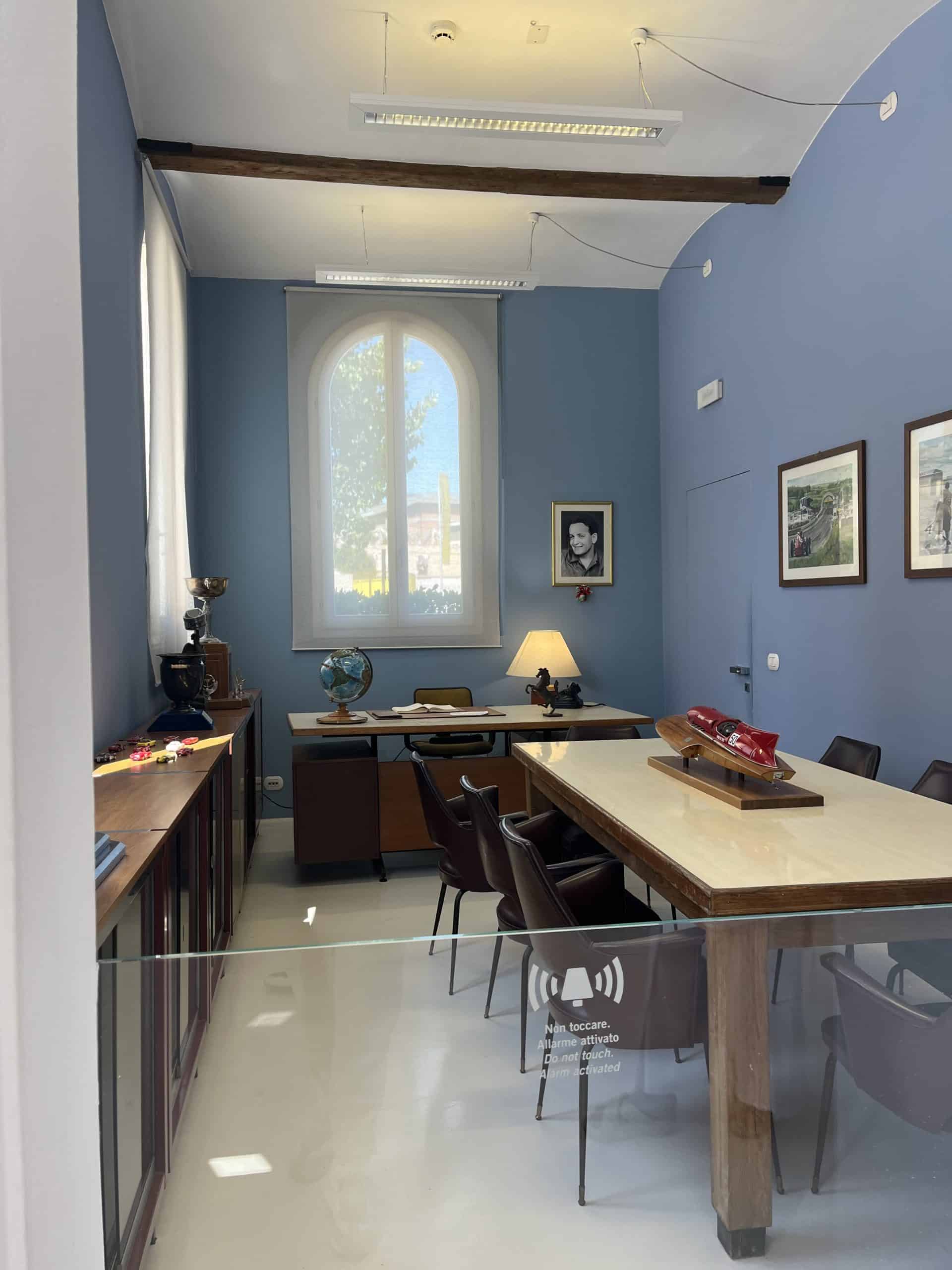 Office with wooden table, vintage chairs, blue walls, framed photos, and large arched window.