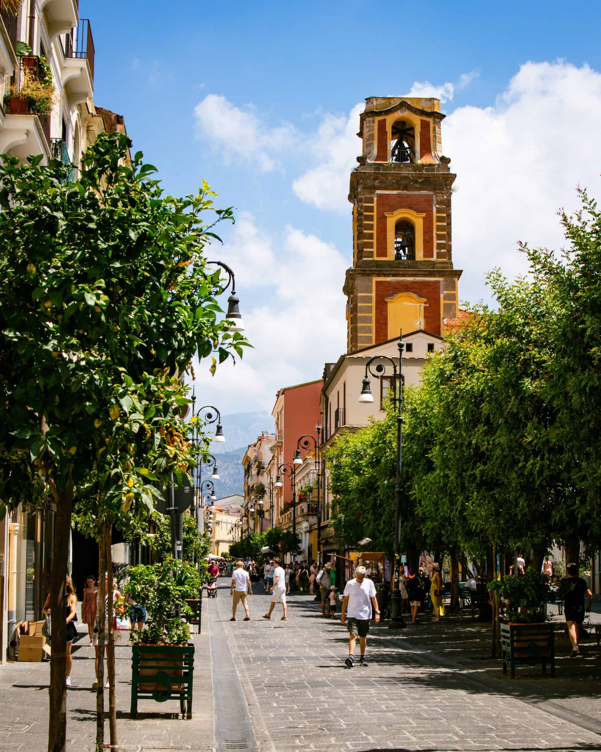 A large tower in the background and a pedestrian walkway lined with trees where people are walking