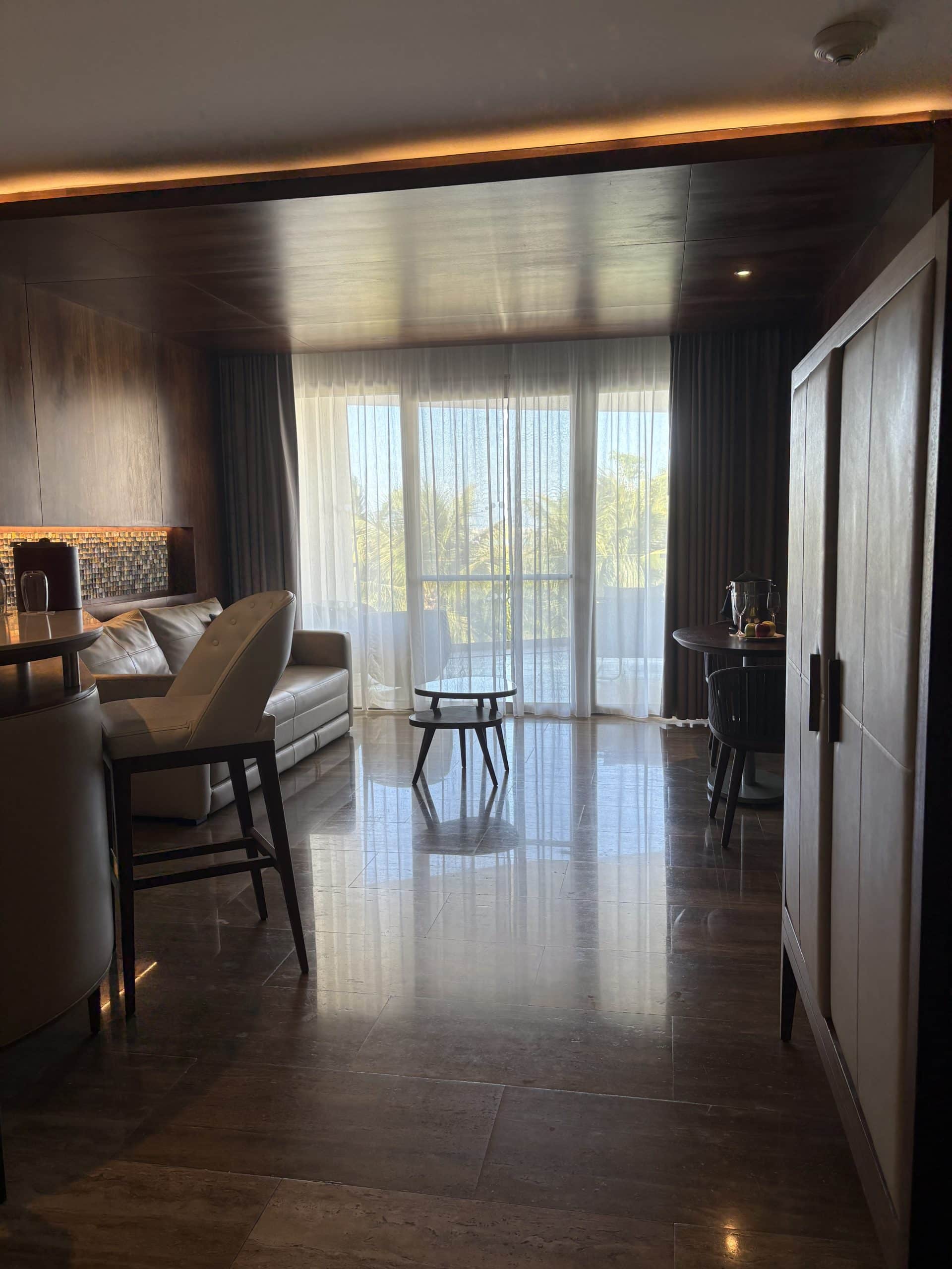 Indoor hotel room with large windows, comfy seating, and scenic views of palm trees and ocean.