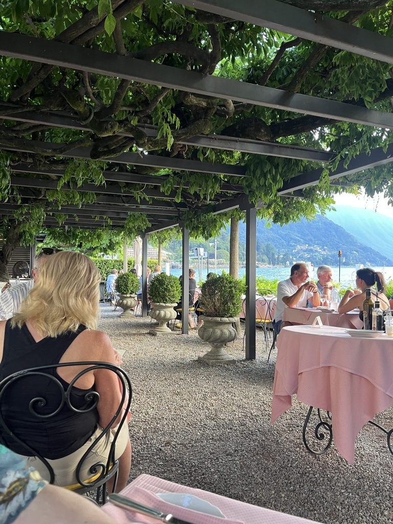 People sitting under a tree covered pergola eating at restaurant tables by the lake